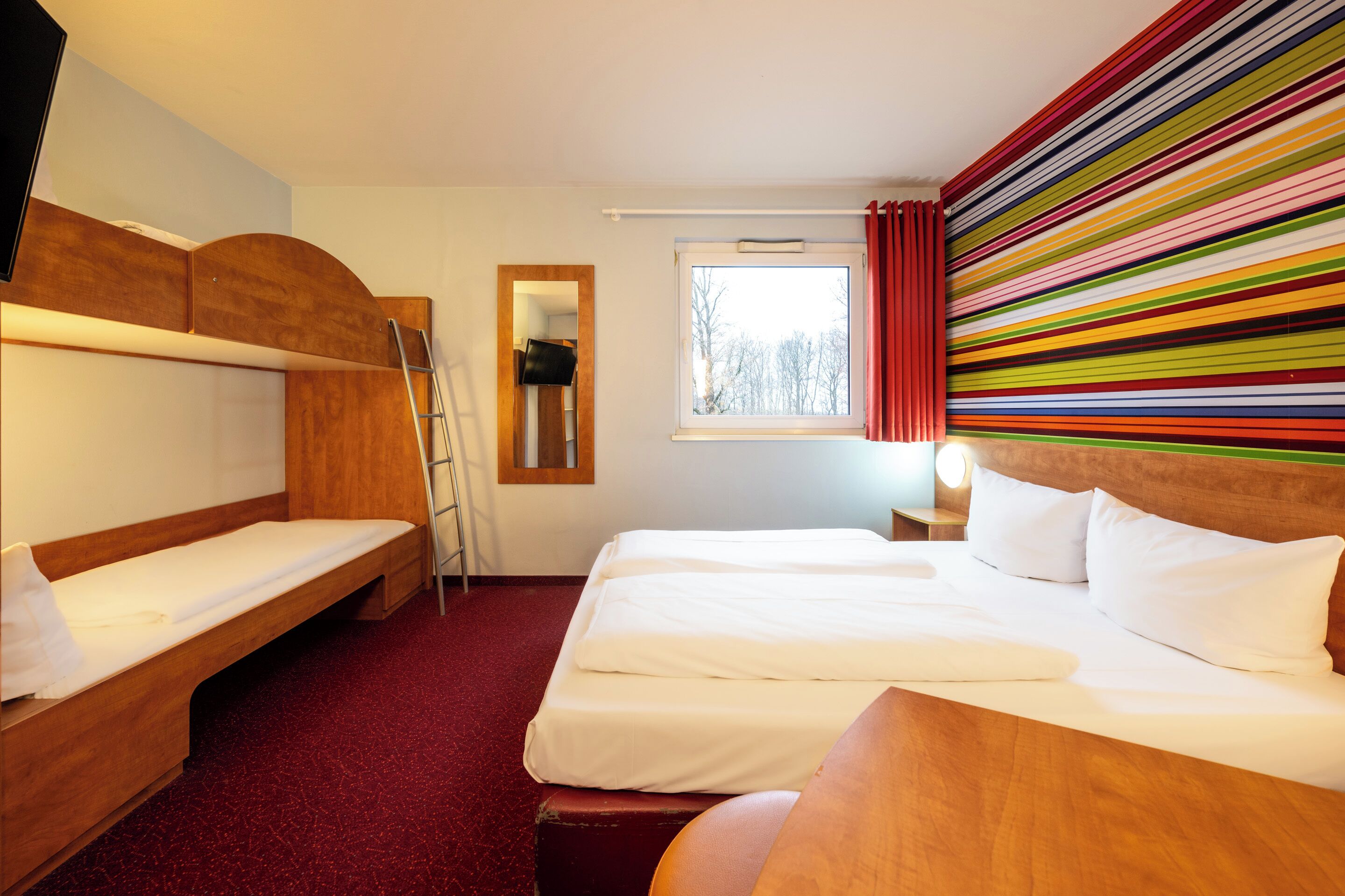 Een hotelkamer met twee bedden, een houten hoofdeinde, een rood tapijt, een raam met gordijnen en een spiegel.
