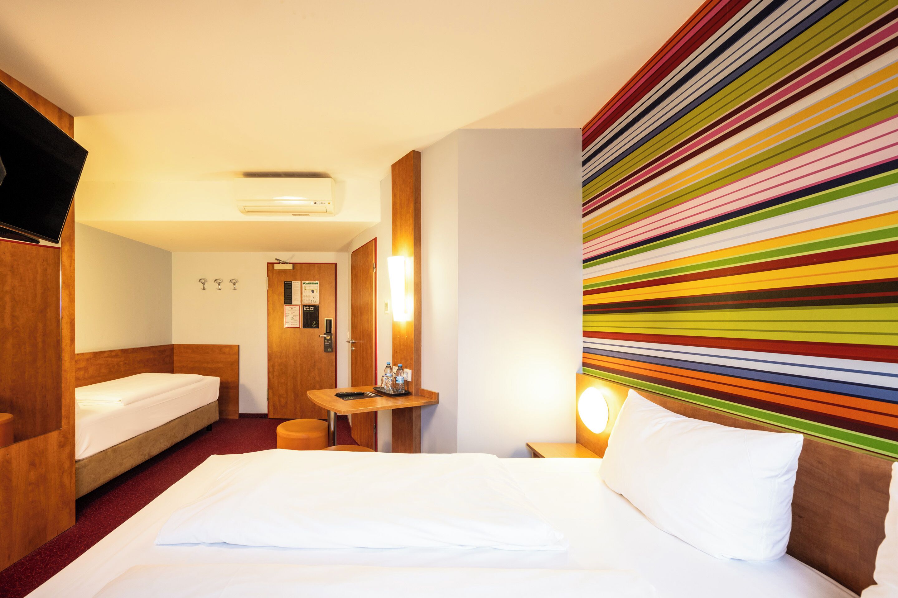 Ein Hotelzimmer mit einem Bett, einem Schreibtisch, einem Sessel und einem Fernseher, sowie einer Wand mit bunten Streifen.