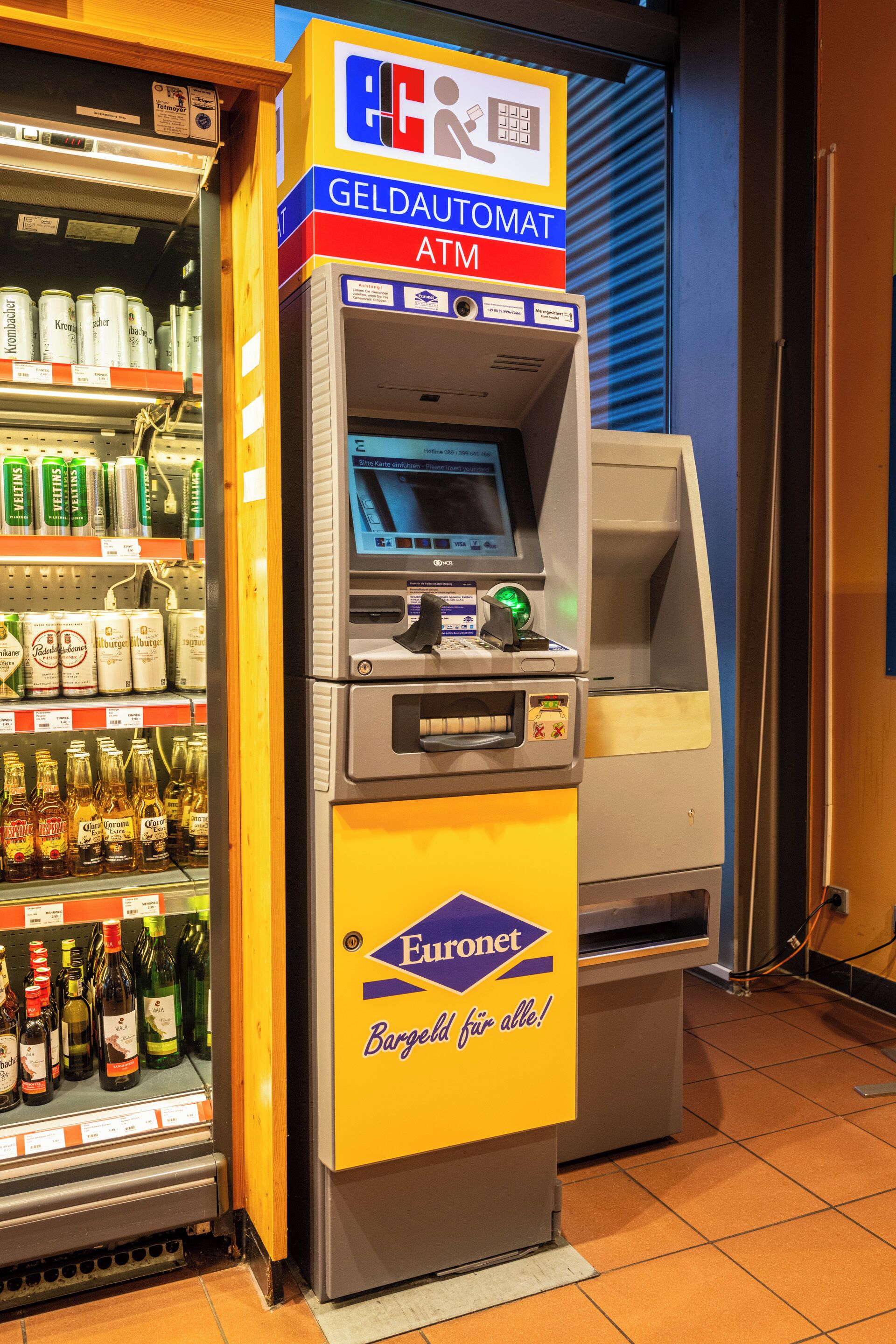 Un distributeur automatique de billets, doté d’une enseigne jaune et bleue, est entouré d’étagères remplies de boissons en bouteille.