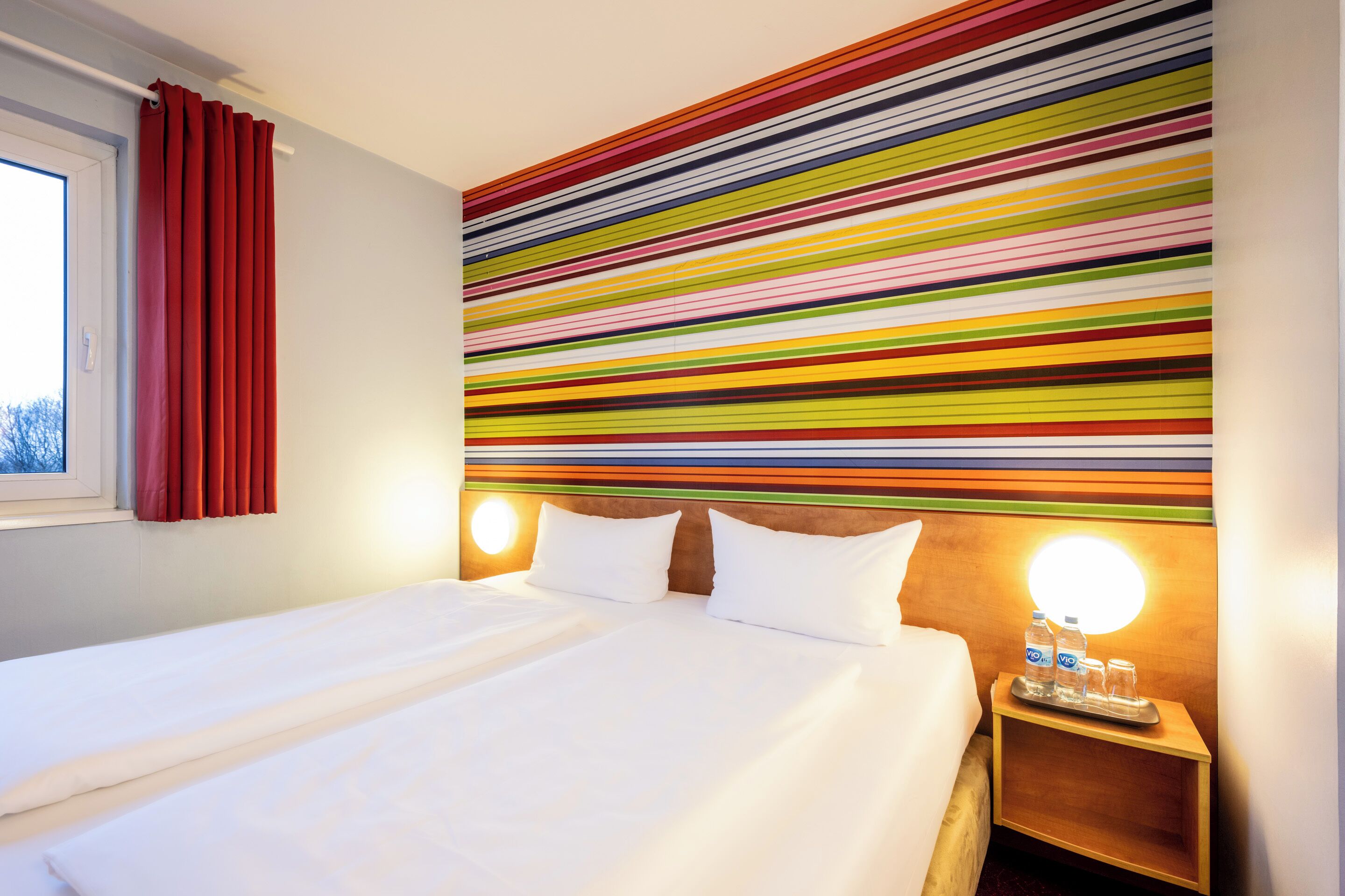 A hotel room with a large bed, a headboard with a colorful striped design, a nightstand with a lamp, and a window with red curtains.