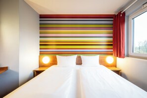 A hotel room with a large bed, two bedside tables, a colorful striped headboard, and a window with curtains.
