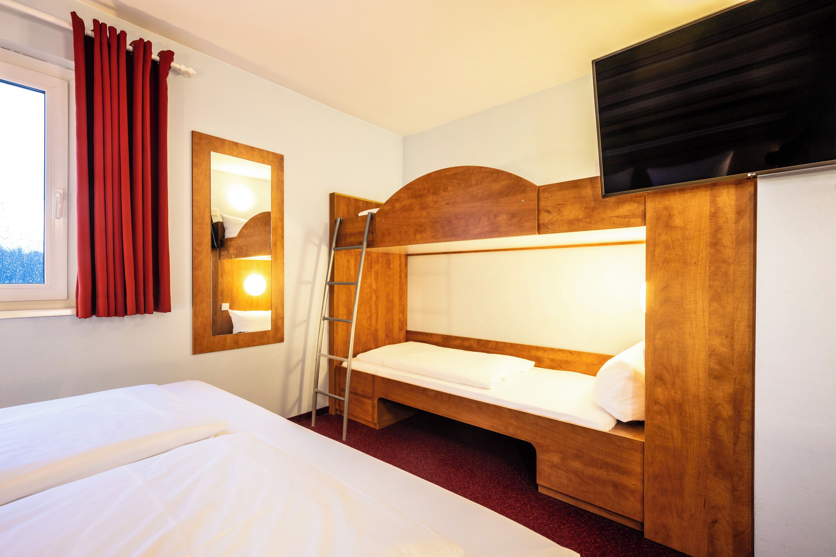 A hotel room with a single bed, a wooden headboard, a small desk, a mirror, and a TV mounted above the bed.