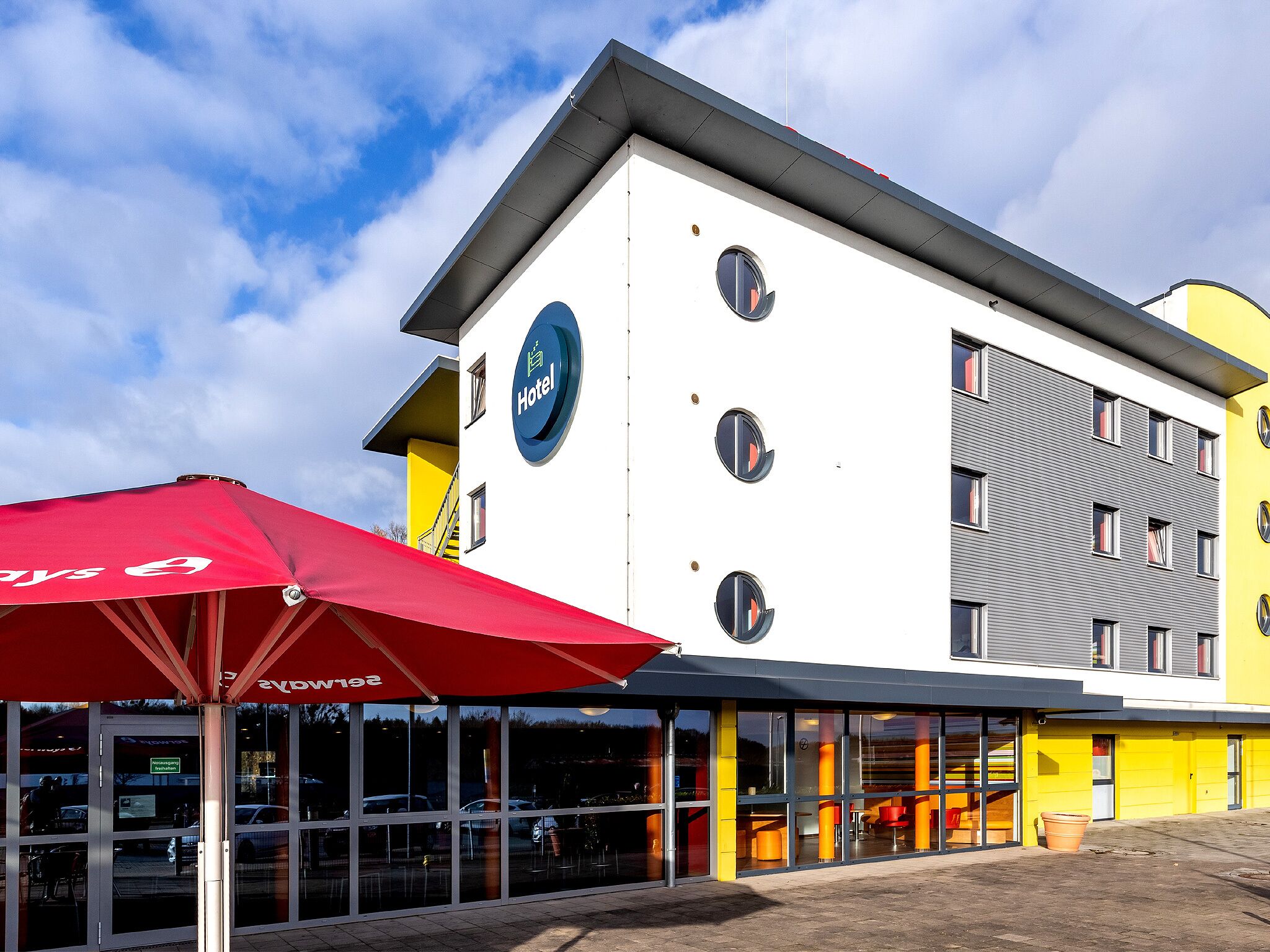 A modern hotel with a prominent 'Hotel' sign, large windows, and a red umbrella.