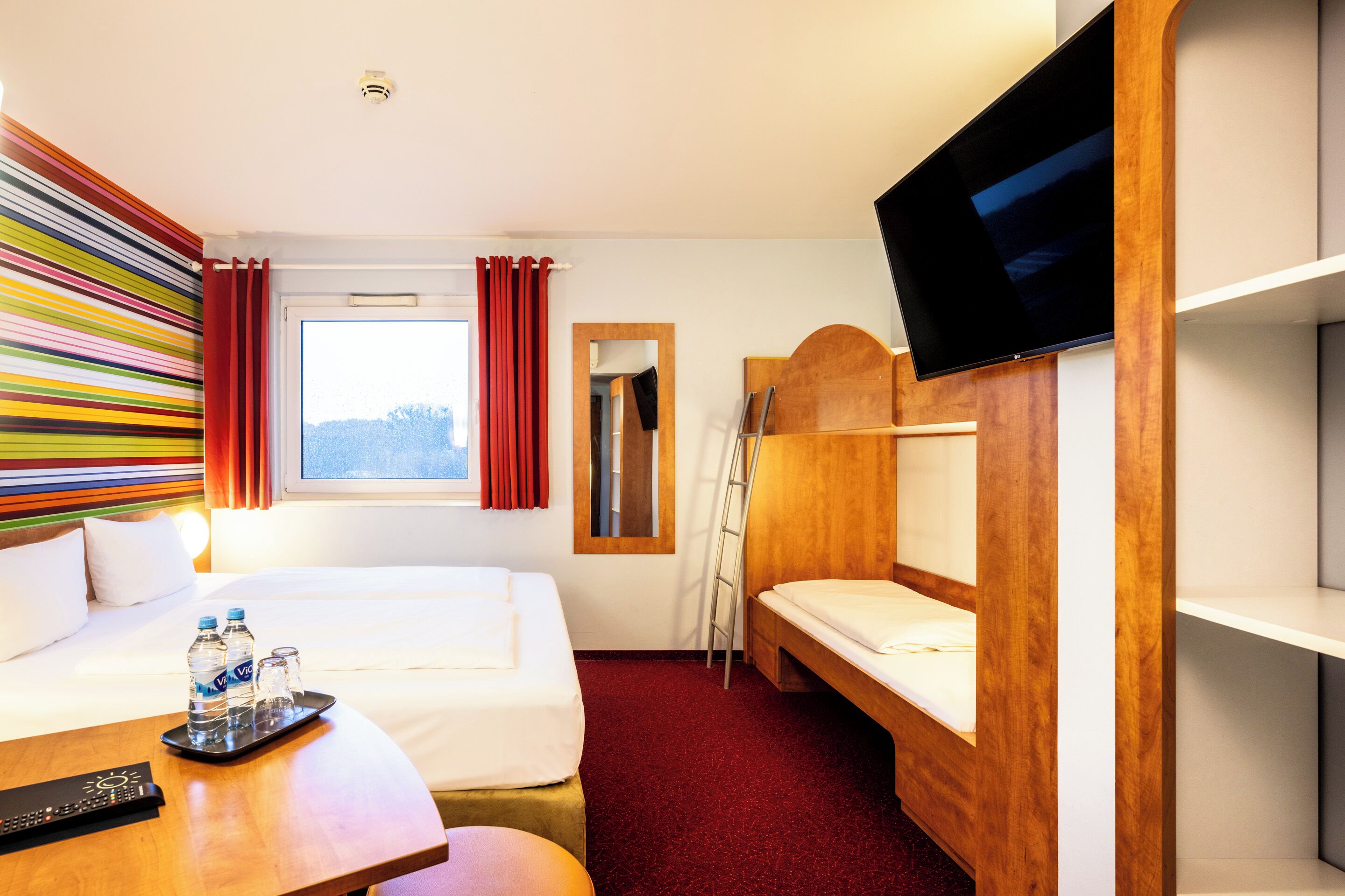 A hotel room with a bed, a desk with a tray of water bottles, a TV, a window with curtains, and a bunk bed.