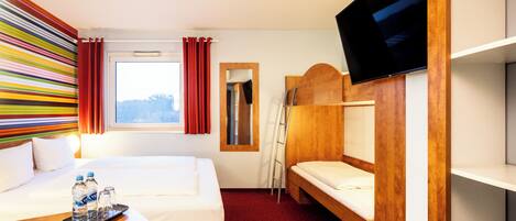 A hotel room with a bed, a desk with a tray of water bottles, a TV, a window with curtains, and a bunk bed.