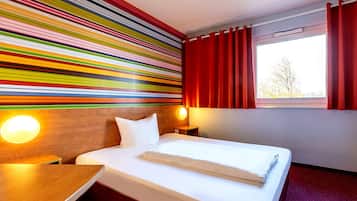 A hotel room with a bed, a wooden headboard, a bedside table, a nightstand, a window with red curtains, and a colorful striped wall.