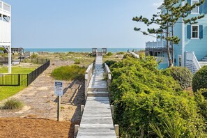 A wooden boardwalk leading to beach chairs, surrounded by greenery and a beach house.