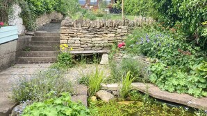 Una escalera de piedra conduce a una estructura de madera con techo de paja, rodeada de abundante vegetación y una pequeña fuente.