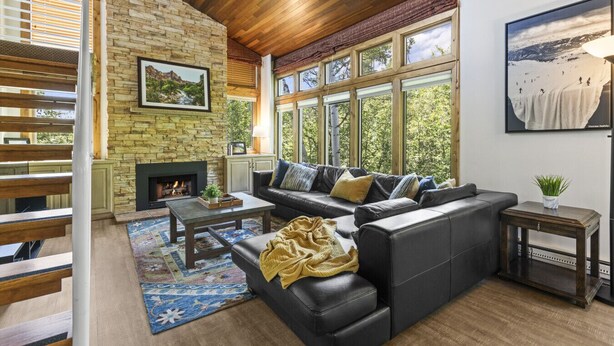 Una sala de estar con pared de piedra, chimenea, sofá de cuero negro y ventanas amplias con vistas a la vegetación.