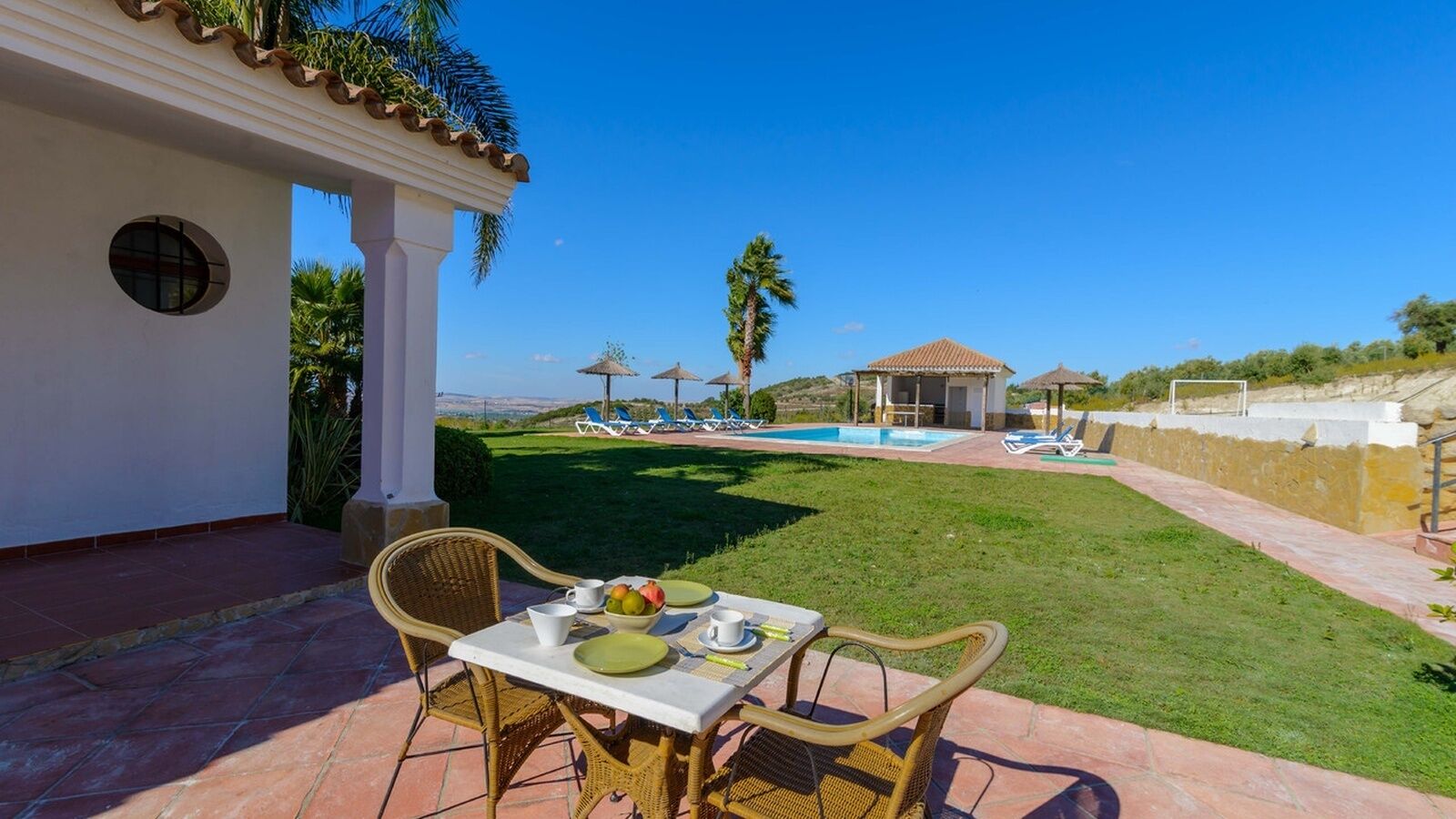 Un patio con una mesa para dos, una piscina y sillones de descanso al fondo.