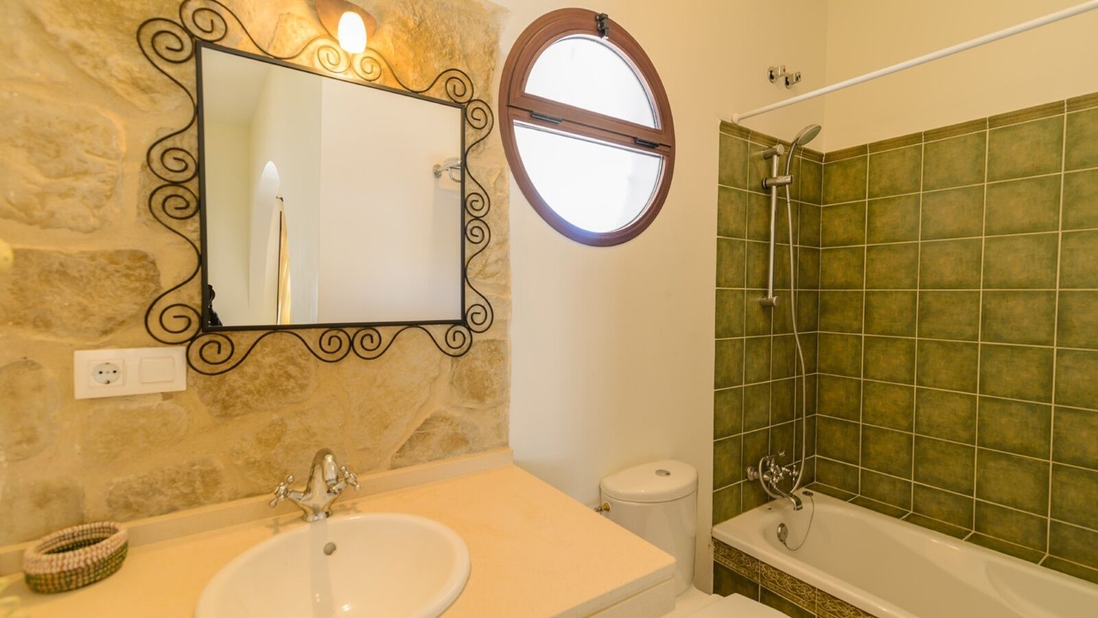 A bathroom with a stone wall, a round mirror, a sink, a toilet, and a bathtub with green tiles.