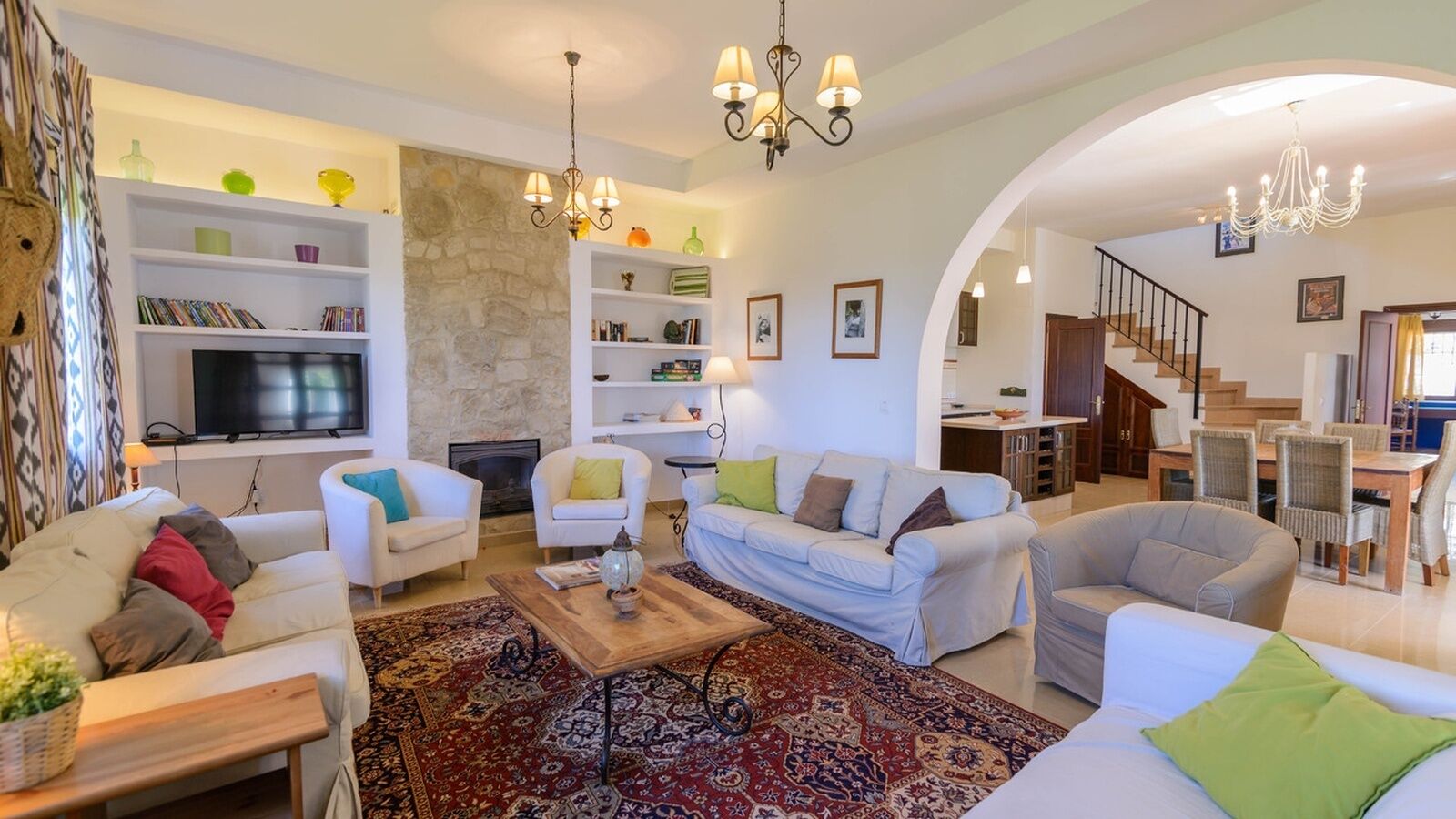 Amplia sala de estar con sofás blancos, una mesa de centro y una chimenea. Hay estanterías con libros y objetos decorativos, y una escalera que conduce a otro nivel.