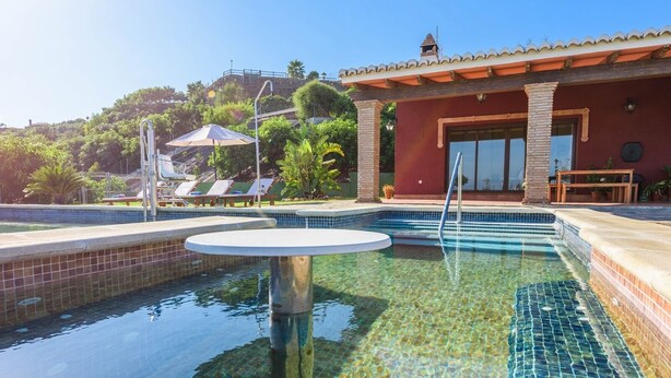 Una piscina con borde de azulejos, un área de descanso con sillas y una sombrilla, y un edificio con techo de tejas y paredes de ladrillo.