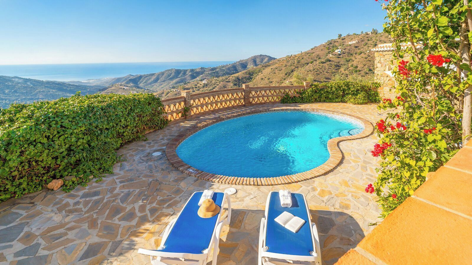A pool with two sun loungers, surrounded by stone flooring and greenery.