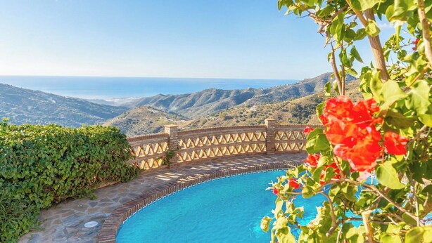 A swimming pool with a view of the sea and hills, surrounded by greenery and a wooden fence.