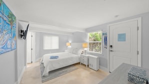 A bedroom with a bed, bedside tables, a nightstand, a window with blinds, a door, and a painting on the wall.