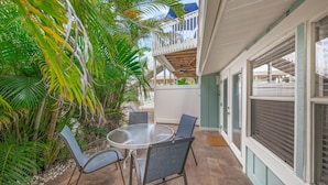 An outdoor patio with a round glass table and four chairs, surrounded by lush greenery and a view of a building with a balcony.