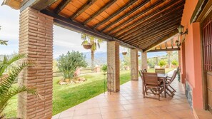 A covered patio with a brick column, a table with chairs, and a view of a landscaped garden with palm trees. - El Pino de Sayalonga by Ruralidays (Málaga)