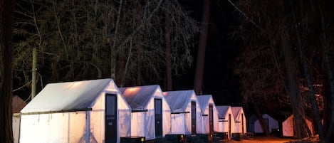 A row of white tents with a single door and a window, set in a forested area at night.
