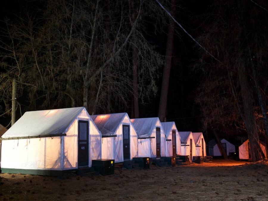 A row of white tents with a single door and a window, set in a forested area at night.