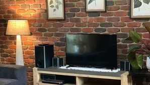 A living room with a brick wall, a flat-screen TV on a wooden stand, a lamp, and framed botanical prints. - Island getaway near farmland, a bird sanctuary, nature walks, & close  to beach (Comerong Island)