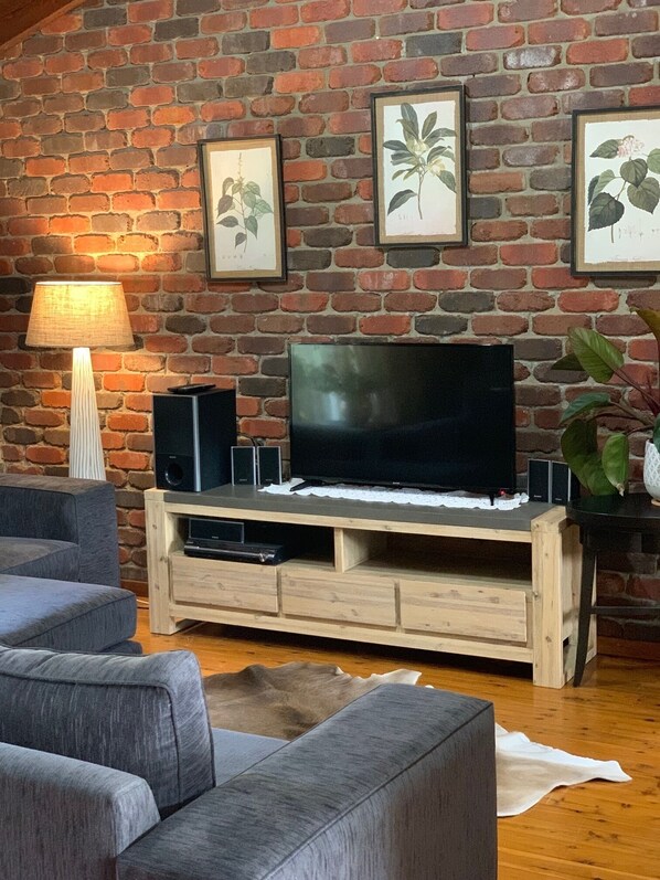 A living room with a brick wall, a flat-screen TV on a wooden stand, a lamp, and framed botanical prints.