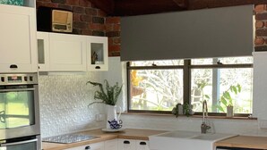A kitchen with white cabinetry, a wooden island, two white chairs, a potted plant, and a window with a view of greenery. - Island getaway near farmland, a bird sanctuary, nature walks, & close  to beach (Comerong Island)