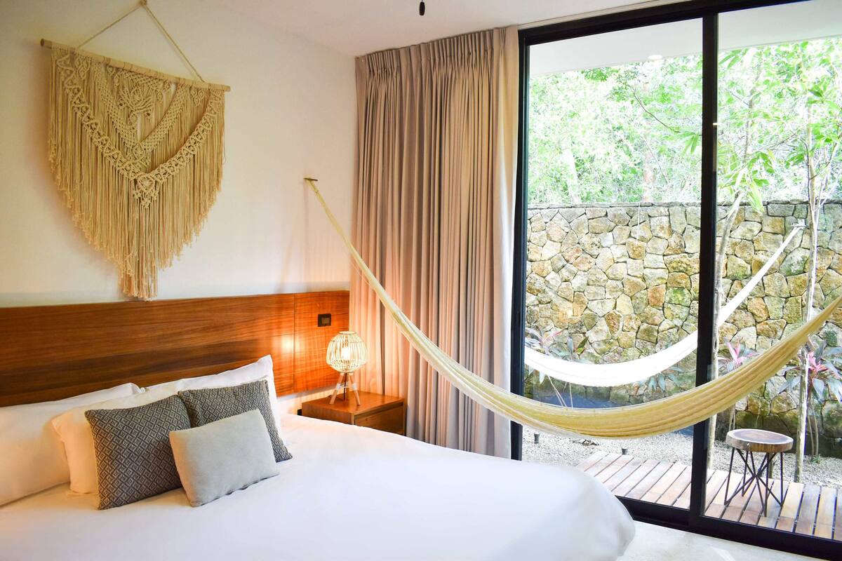 Une chambre d’hôtel avec un lit, une décoration murale en macramé, une table de chevet avec une lampe, et une vue sur un mur de pierre et de la verdure, le tout visible à travers une grande fenêtre.