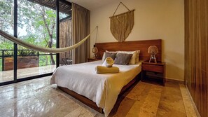 A bedroom with a large bed, a nightstand, a lamp, and a view of trees through a window.