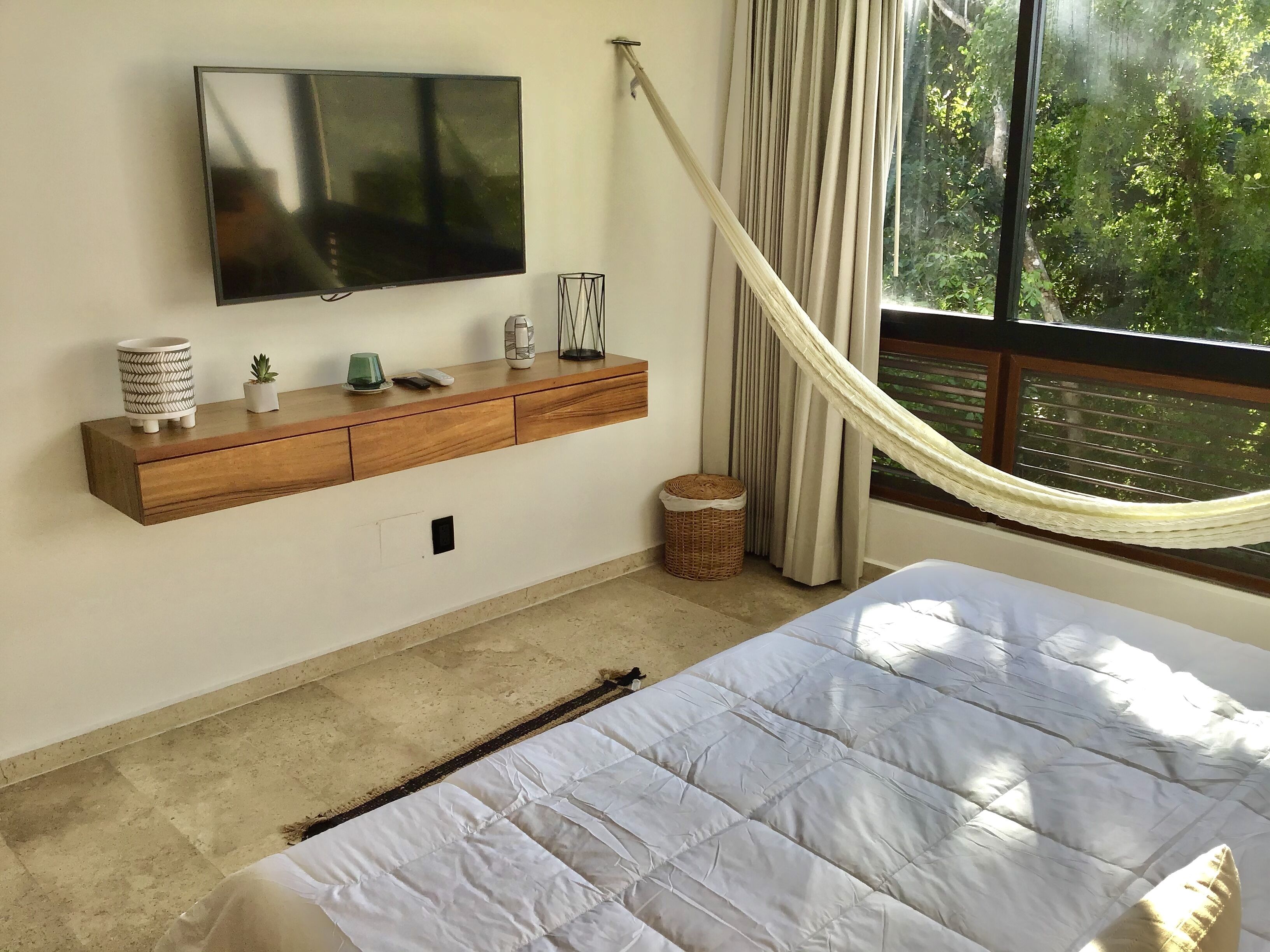 A bedroom with a bed, a TV mounted on the wall, a wooden console table with decorative items, a window with curtains, and a hammock.