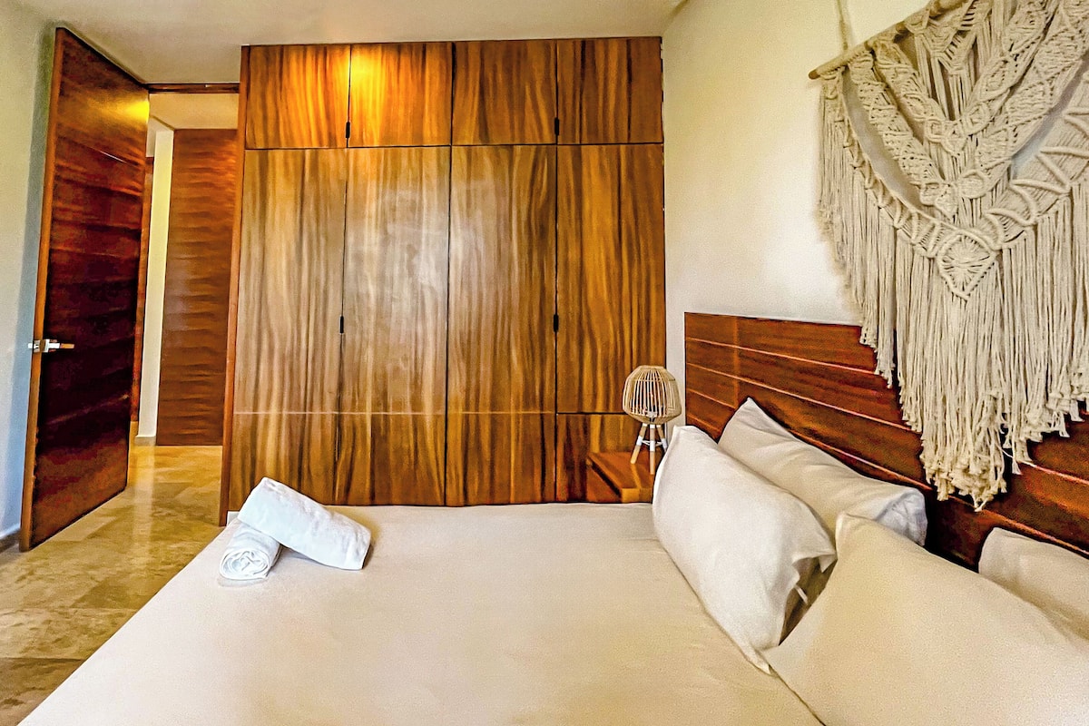 Une chambre d’hôtel avec une armoire en bois, un lit avec du linge de maison blanc et une décoration murale en macramé.