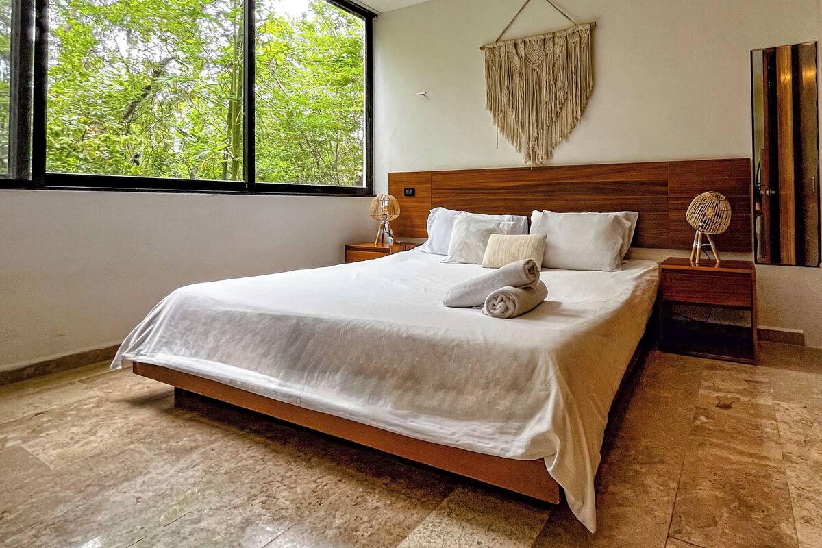 Une chambre avec un grand lit, une tête de lit en bois et une vue sur les arbres par la fenêtre.