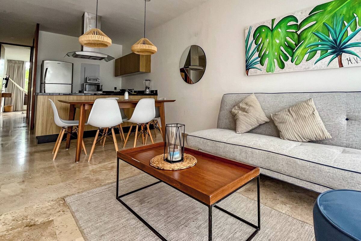 Un salon moderne avec un canapé gris, une table basse en bois et un coin repas comprenant des chaises, avec une cuisine en arrière-plan.