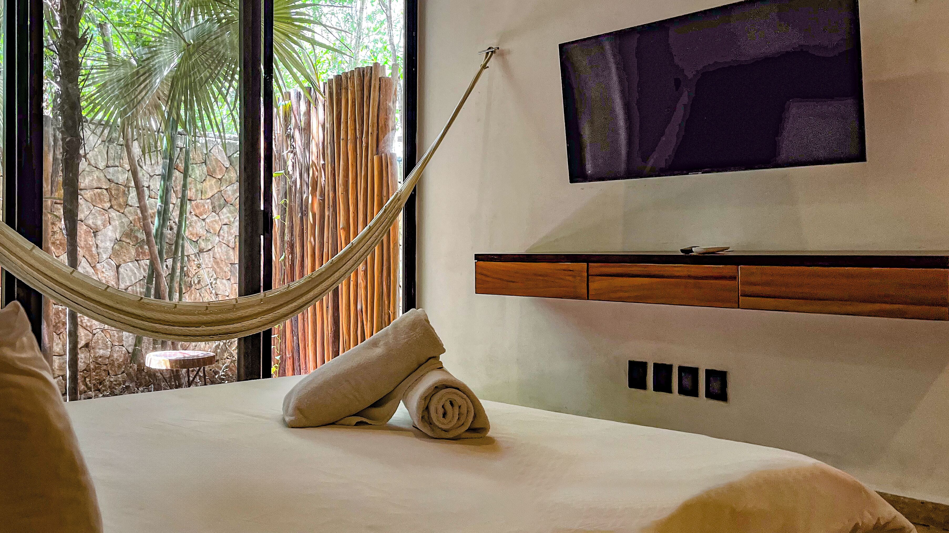 A bedroom with a large window, a hammock, a TV mounted on the wall, and a wooden cabinet.