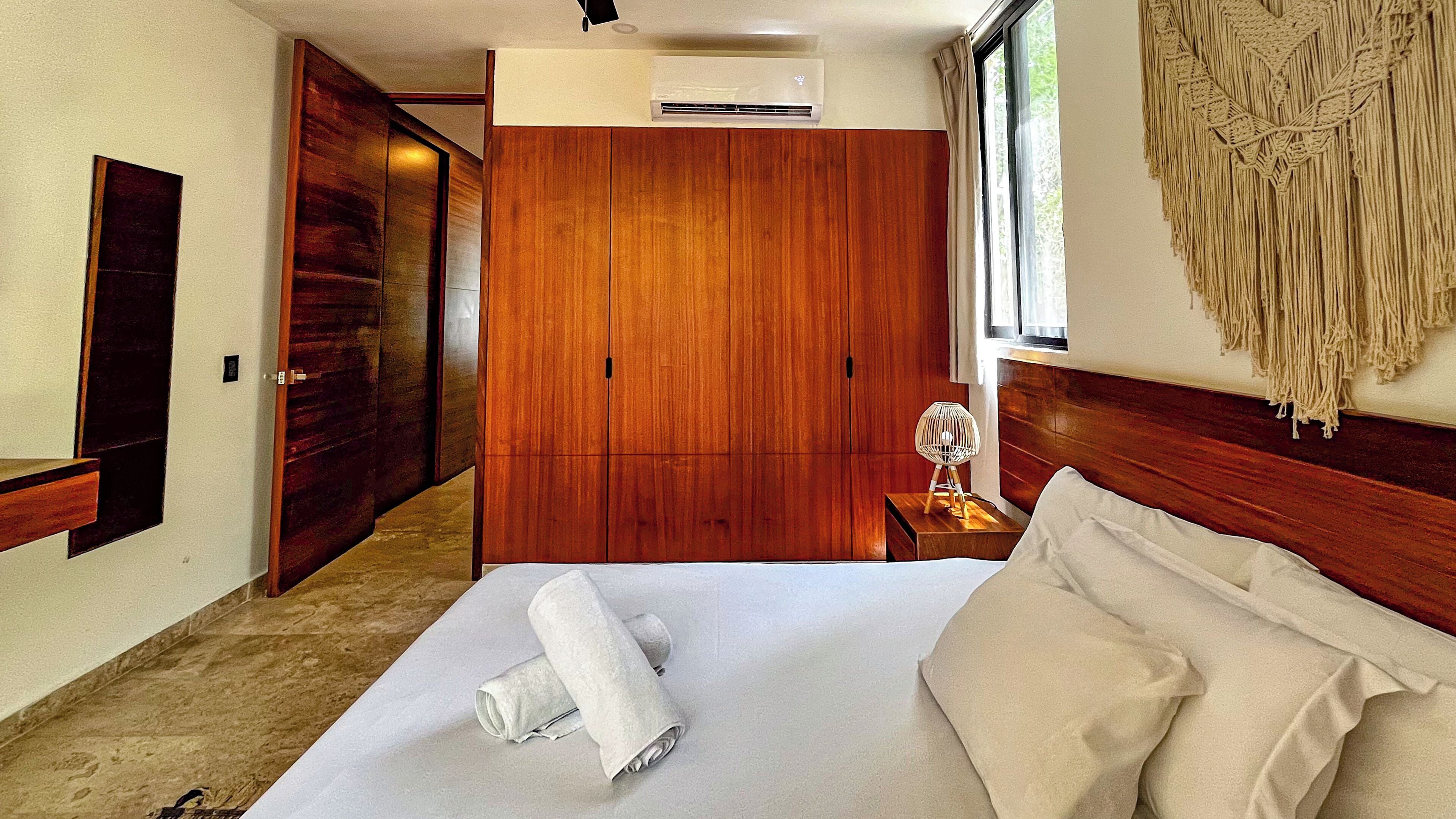 A hotel room with a wooden wardrobe, a bed with white linens, a bedside table with a lamp, and a large window with a view.