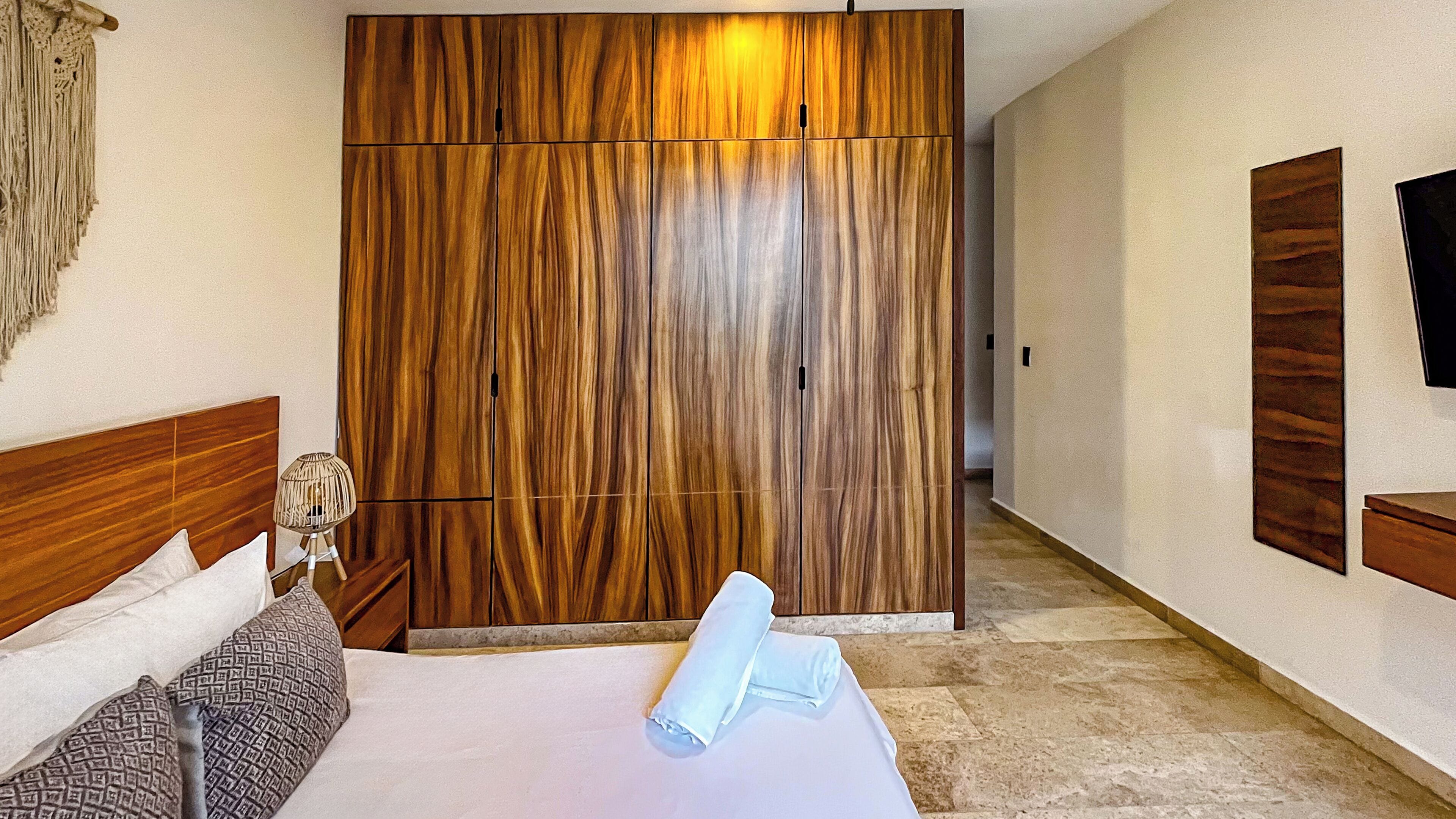 A hotel room with a large wooden wardrobe, a bed with patterned pillows, and a bedside lamp.