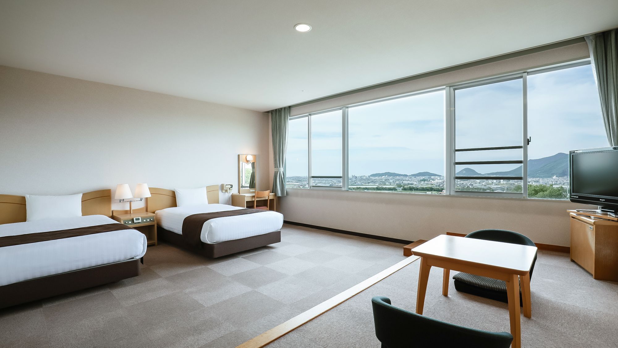 A hotel room with two beds, a desk, a chair, a TV, and a large window with a city view.