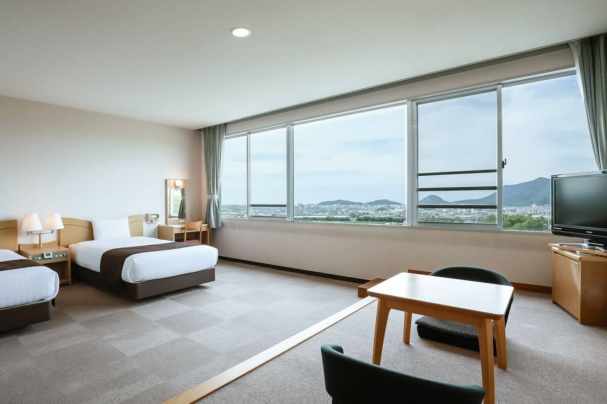 A hotel room with two beds, a desk, a chair, a TV, and a large window with a city view.