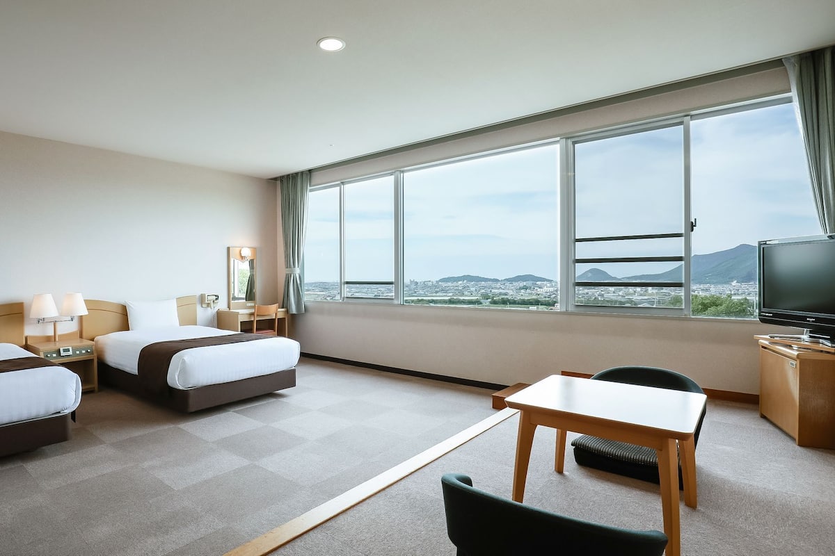 Une chambre d’hôtel avec deux lits, un bureau, une chaise, une télévision et une grande fenêtre donnant sur la ville.