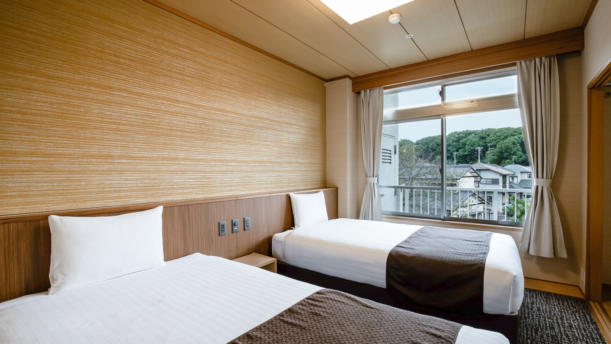 A hotel room with two beds, a wooden headboard, a large window with curtains, and a view of buildings outside.