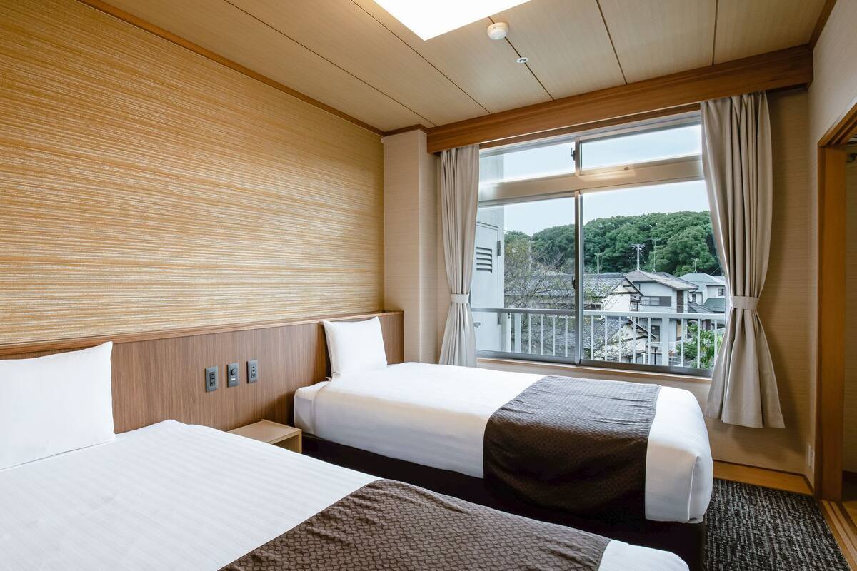 A hotel room with two beds, a wooden headboard, a large window with curtains, and a view of buildings outside.
