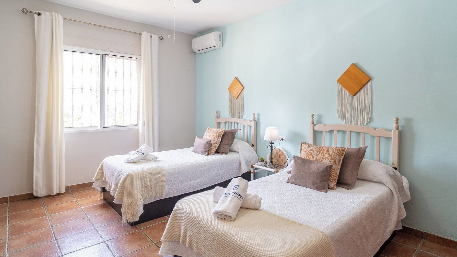 Habitación de hotel con dos camas, una ventana con cortinas, un equipo de aire acondicionado y un adorno de macramé colgado en la pared.