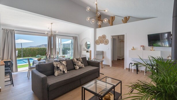 A spacious living area with a sofa, coffee table, and a dining area with chairs. There is a pool visible through large windows, and a flat-screen TV mounted on the wall. - Cortijo Entre Pueblos Cómpeta by Ruralidays (Málaga)