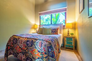 Un dormitorio con una cama grande, una ventana con vistas a follaje y una mesita de noche con lámpara.