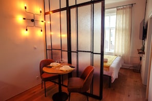 A room with a bed, a dining table with chairs, a wall-mounted light fixture, and a window with curtains. - CAMŌ ROOMS - Boutique Aparthotel (Prague)