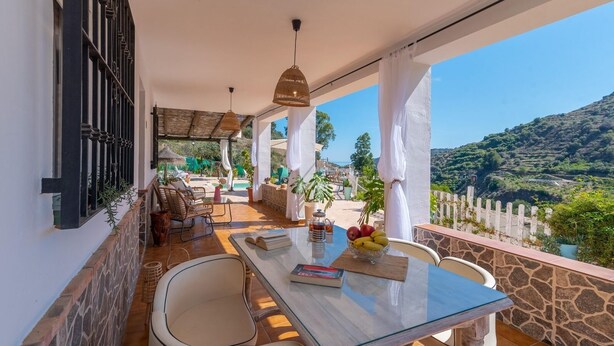 A balcony with a table set for dining, surrounded by wicker furniture, and overlooking a scenic landscape.