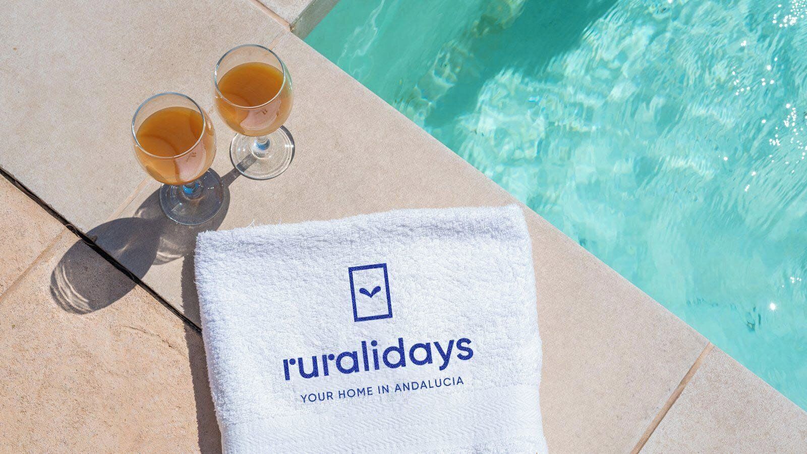 A poolside setting with a towel displaying 'ruralidays' and two glasses of amber liquid.
