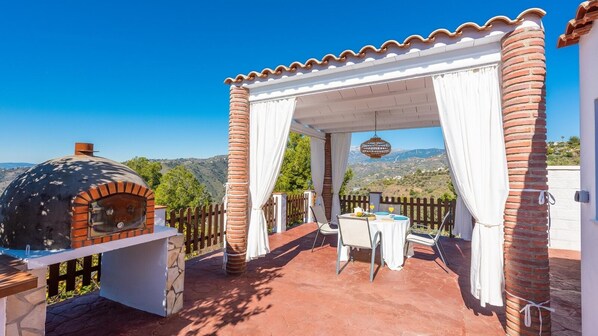 A patio with a brick oven, white curtains, and a table set for two. - Villa Francisco Torrox by Ruralidays (Málaga)