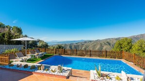 A swimming pool with a wooden fence, lounge chairs, and a view of hills.