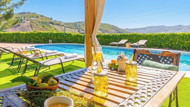 A poolside area with lounge chairs, a wooden table with drinks, and a view of a pool and hills. - La Noria de Iznate by Ruralidays (Málaga)